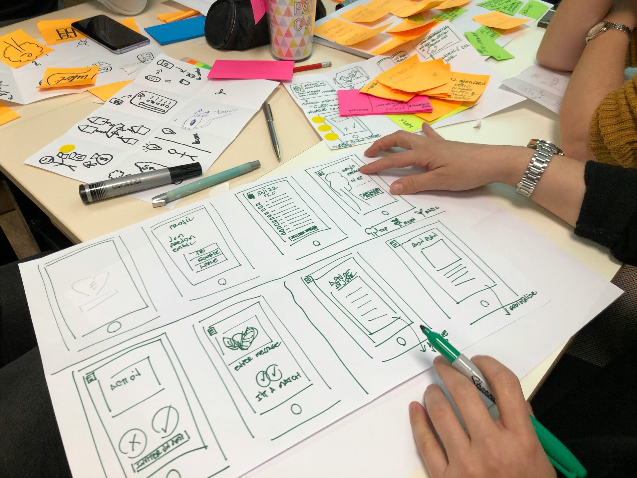Hand-drawn wireframes are on a table with a person contininuing to work on them with a marker in hand.